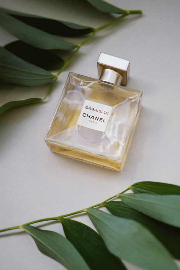 A glass bottle of Chanel perfume on a white table surrounded by green leaves